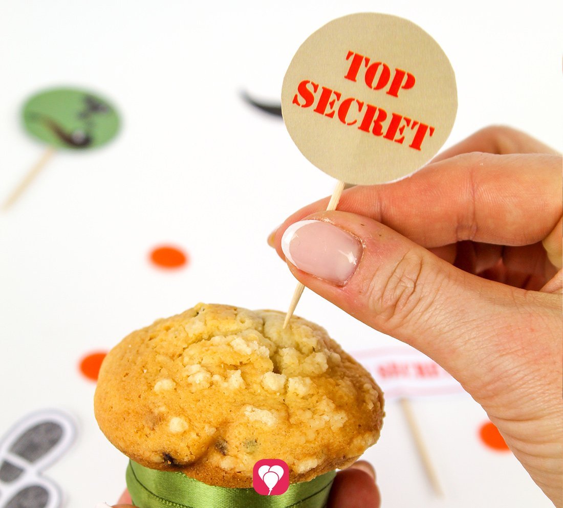Eine Person steckt einen balloonas Detektiv Deko Picker mit der Aufschrift "TOP SECRET" in einen Muffin. 